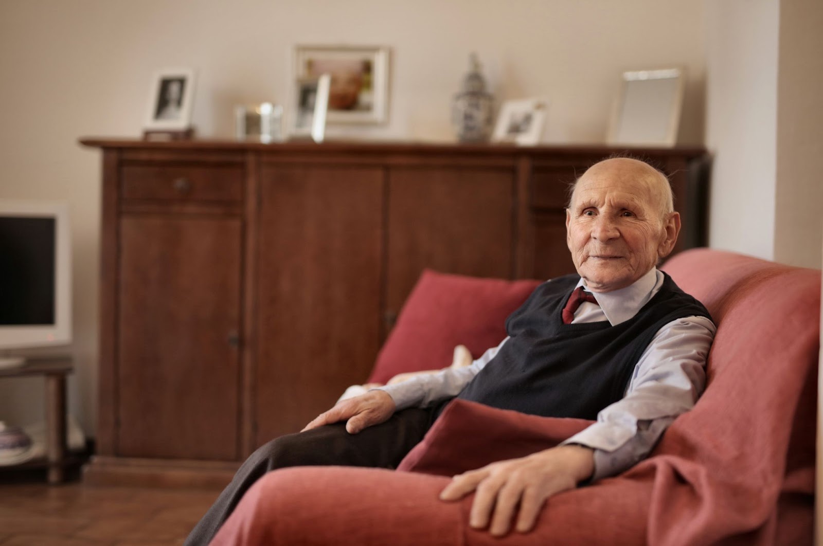 An elderly man in a sweater vest and neat shirt sits alone on a couch in a living room