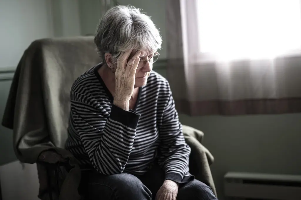 An old woman suffering nursing home abuse in Michigan, sitting in a chair and holding her head.