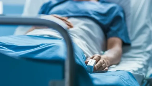 A victim of a brain injury lying down on a bed with a heart monitor on their hand, suffering a vegetative state.