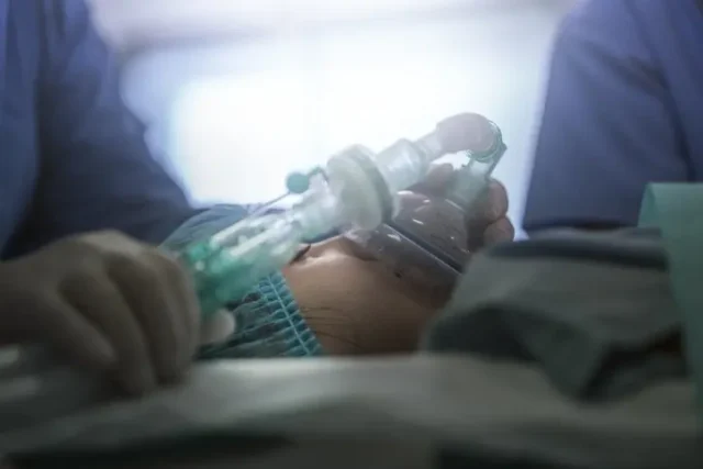 A woman in a coma with an oxygen mask on. Two doctors are putting on the mask on her face.