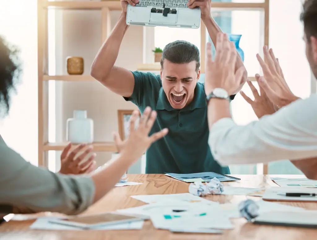 A man screaming and holding a laptop above his head, threatening to throw it down on the table. Someone is assaulted at work.