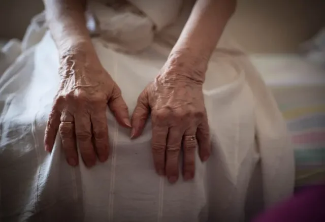 An elderly person with their hand on their knees, sitting on a bed.