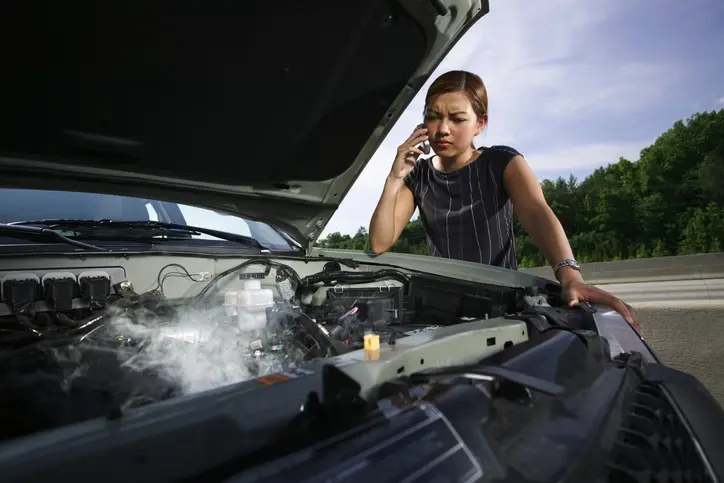A woman calling her insurance for a car accident, staring at a smoking engine.